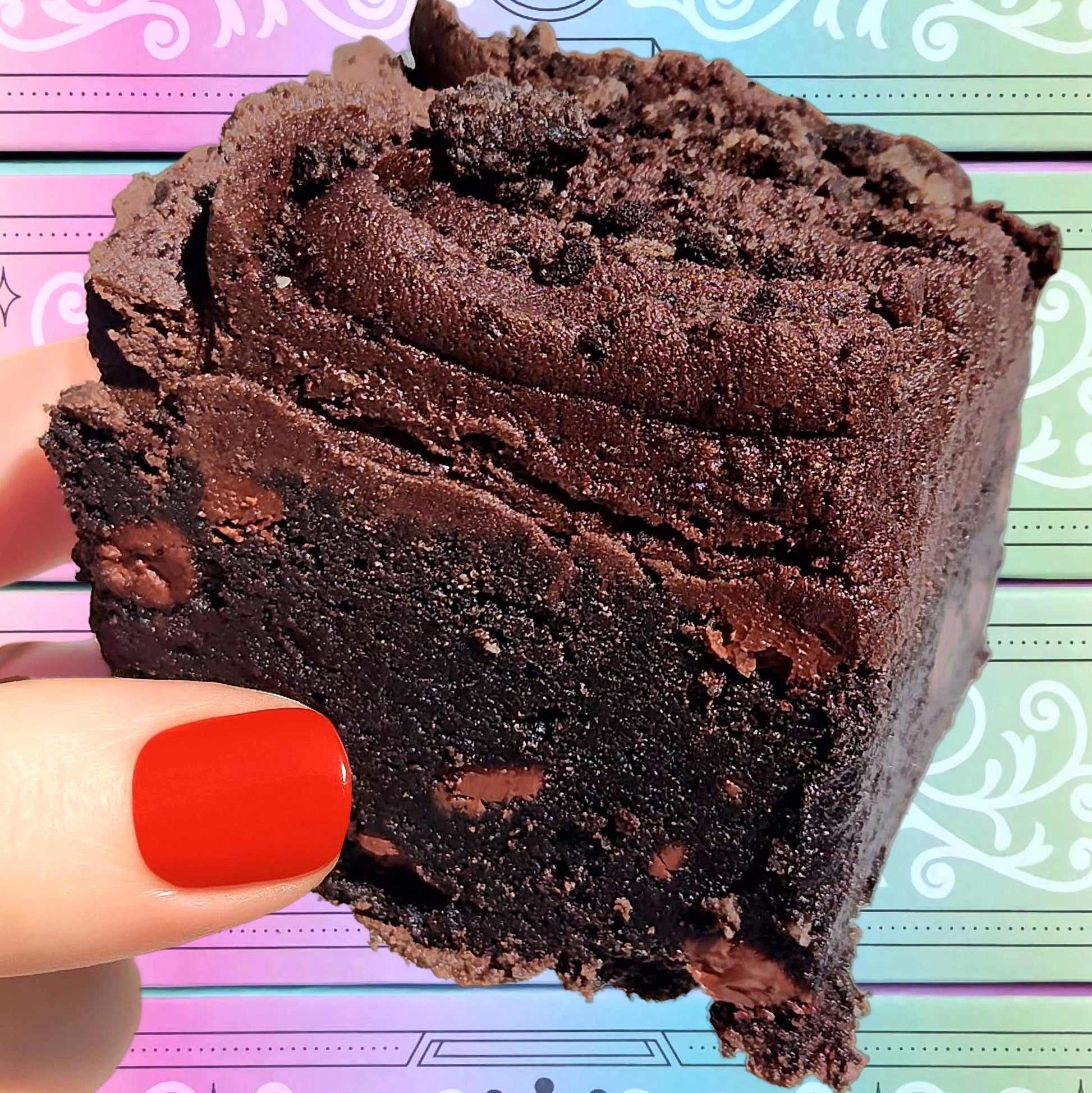 A piece of chocolate stout buttercream shortbread with chocolate chips and Oreo crumbs on top, held by a person with red nails.