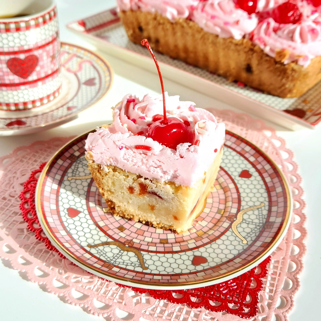 Slice of shortbread with pink cherry frosting and a cherry on top, served on a decorative plate with a heart-themed cup in the background.
