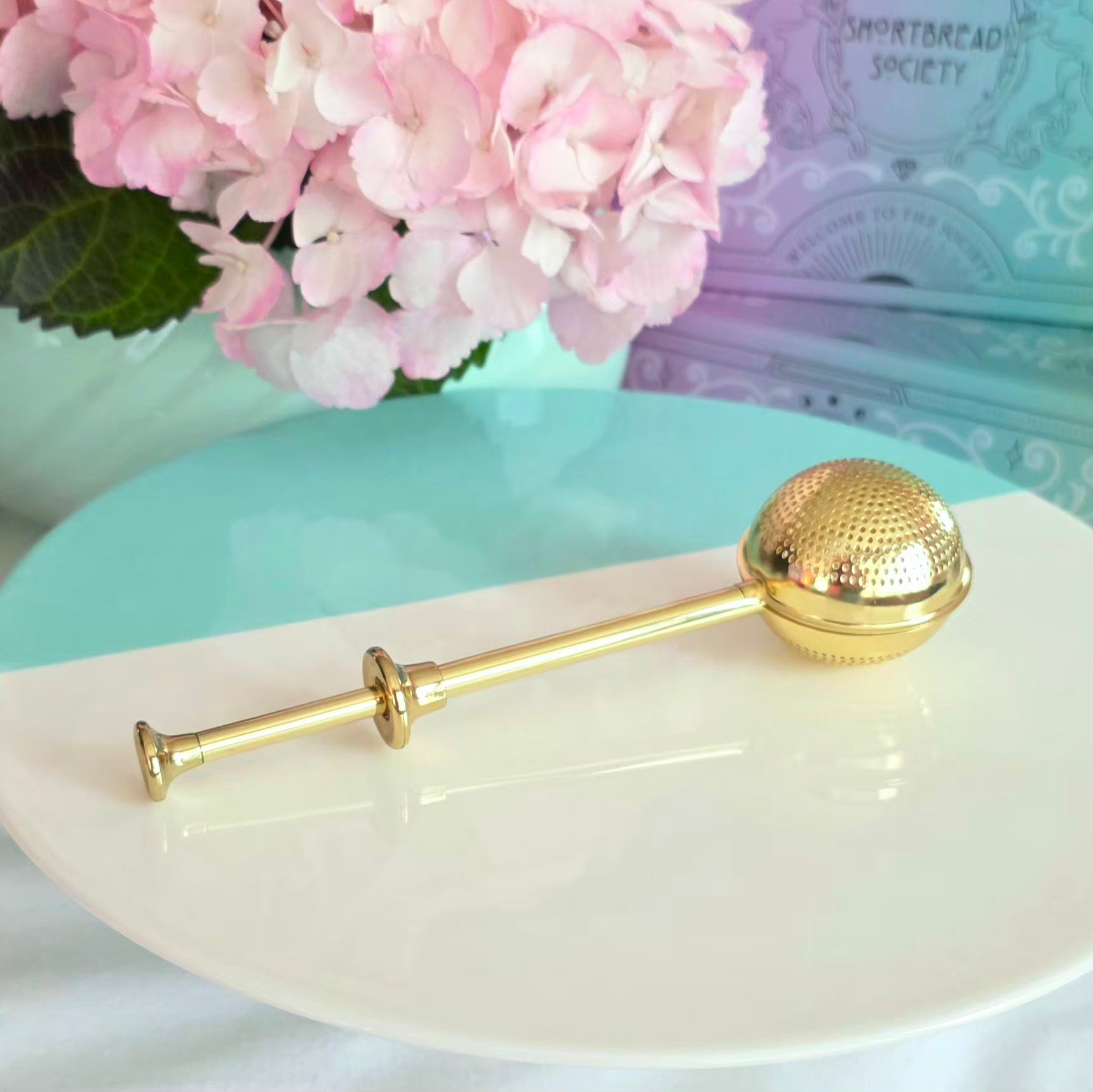 Gold tea ball on a white plate with pink flowers in the background